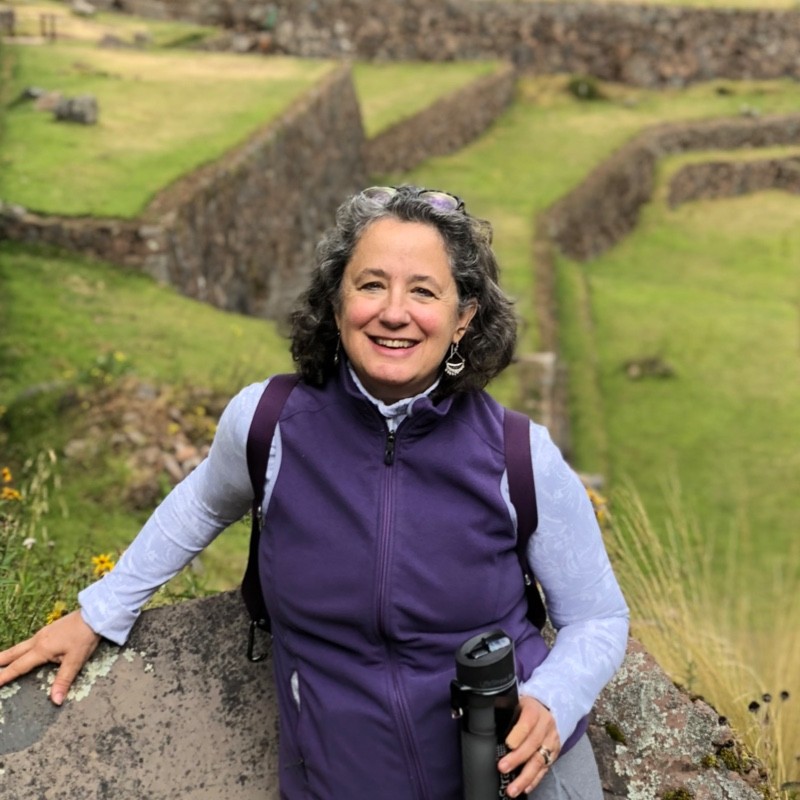 Smiling woman wearing a purple vest, standing among stone terraces outdoors, holding a black water bottle and wearing a backpack.